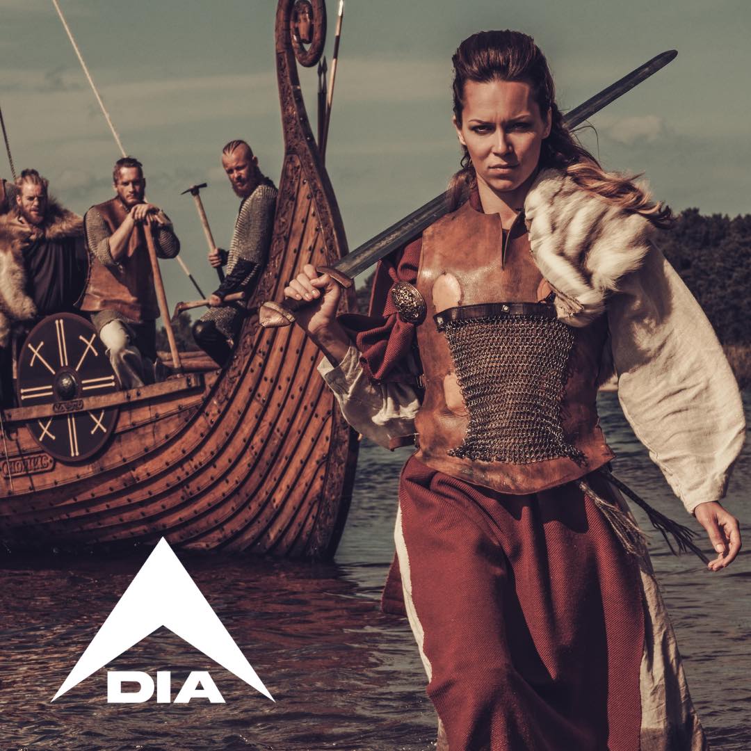 Viking woman holding a sword by a longship with three men, featuring a white DIA logo.