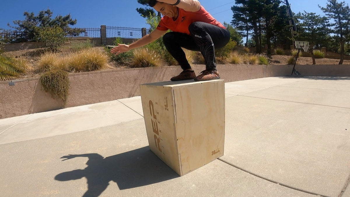 Man outdoors performing box jumps on a wooden box, wearing a red DIA shirt and black athletic leggings.