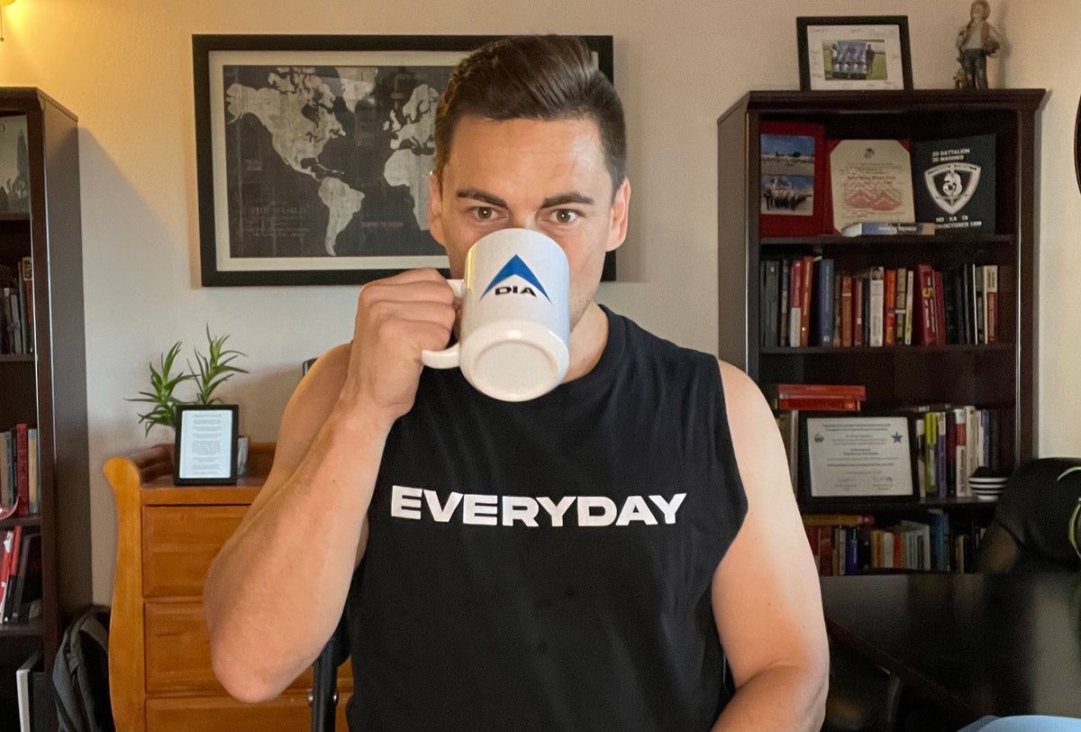 Man in a home office drinking from a white mug with the DIA logo, wearing a black DIA Everyday tank top.