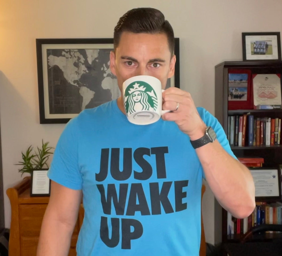 Man in an office drinking from a Starbucks mug, wearing a blue DIA "JUST WAKE UP" t-shirt.