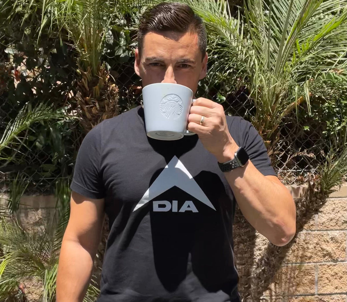 Man standing outdoors drinking from a white Starbucks mug, wearing a black DIA logo t-shirt.