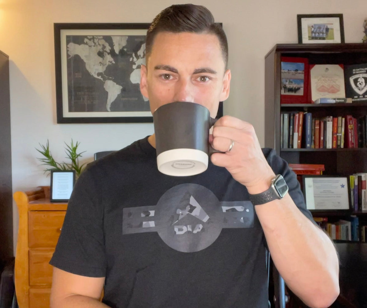 Man in an office drinking from a mug, wearing a black t-shirt with a grey camo DIA logo.