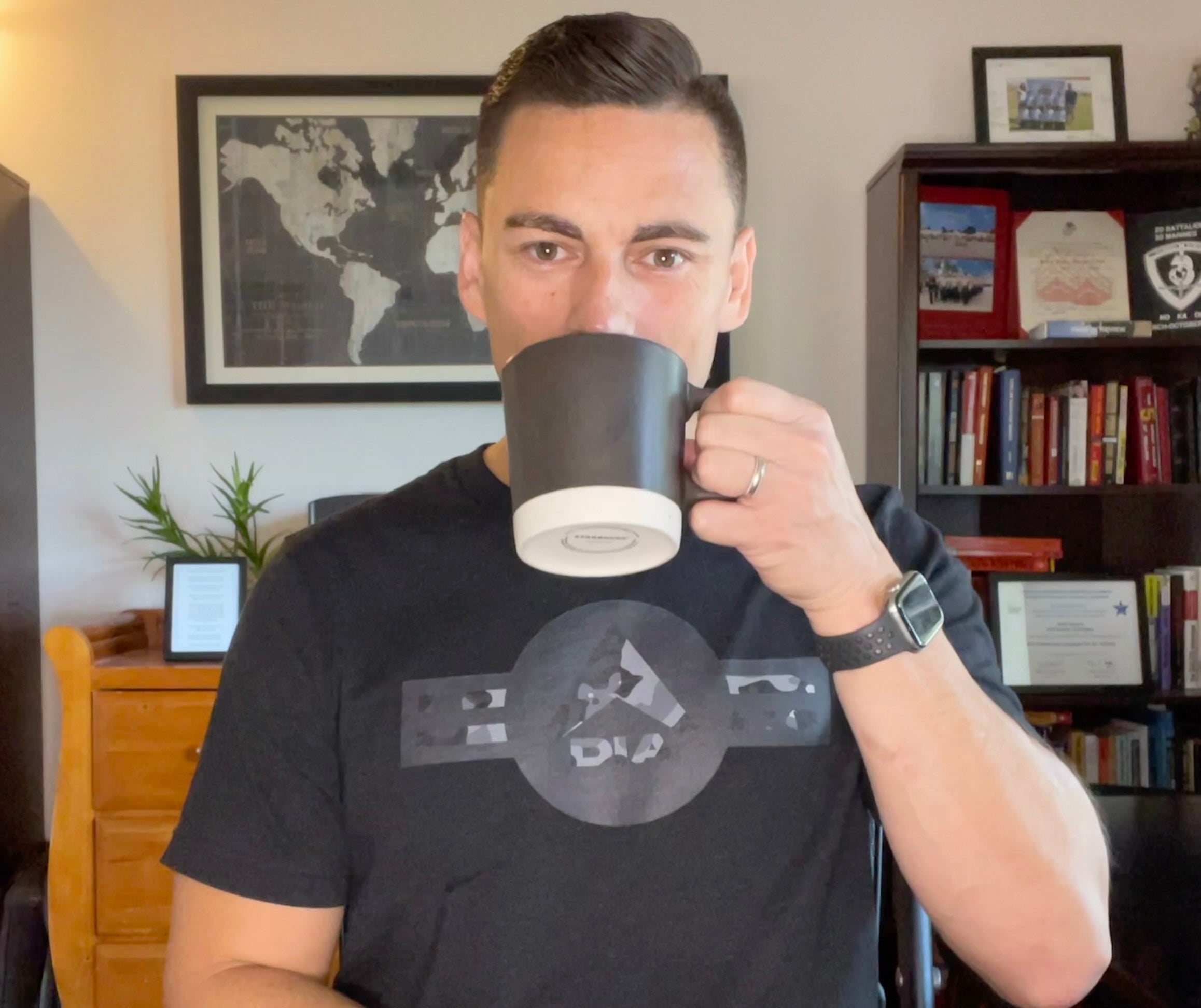Man in an office drinking from a mug, wearing a black t-shirt with a grey camo DIA logo.