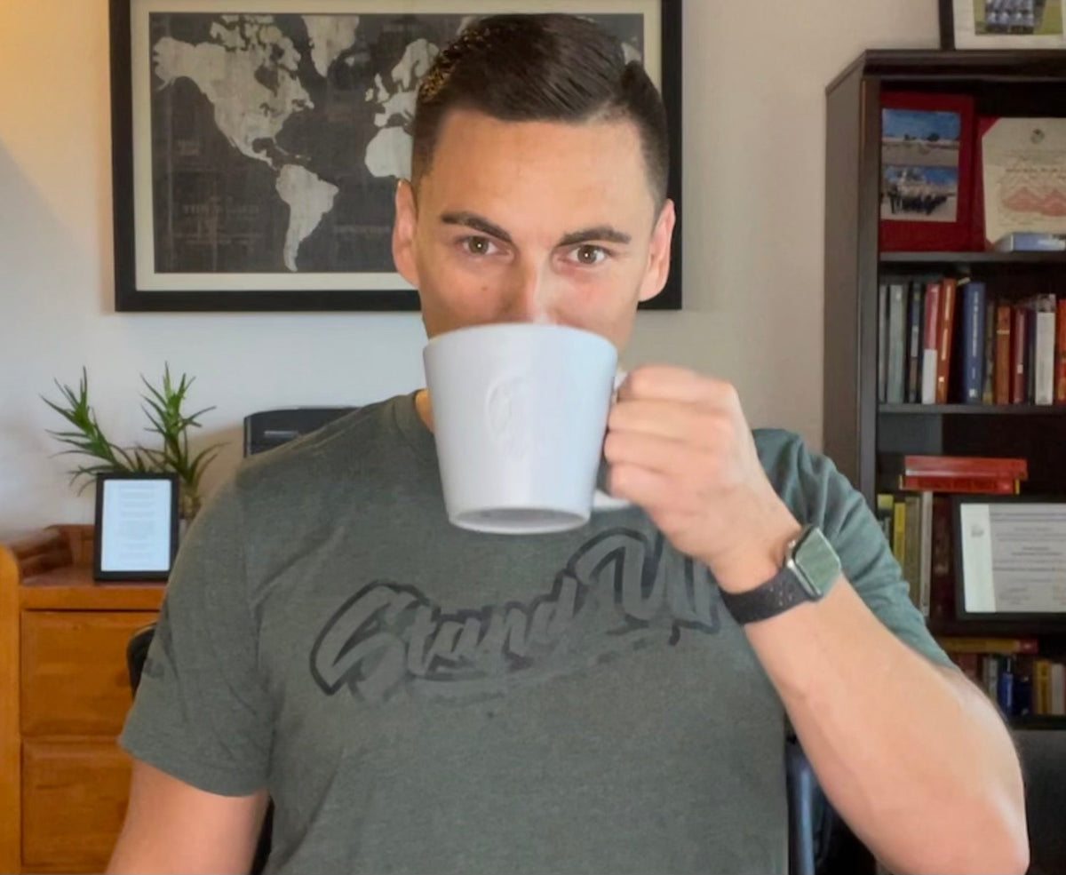 Man in a home office drinking coffee, wearing a grey DIA Stealth Stand Up T-Shirt.