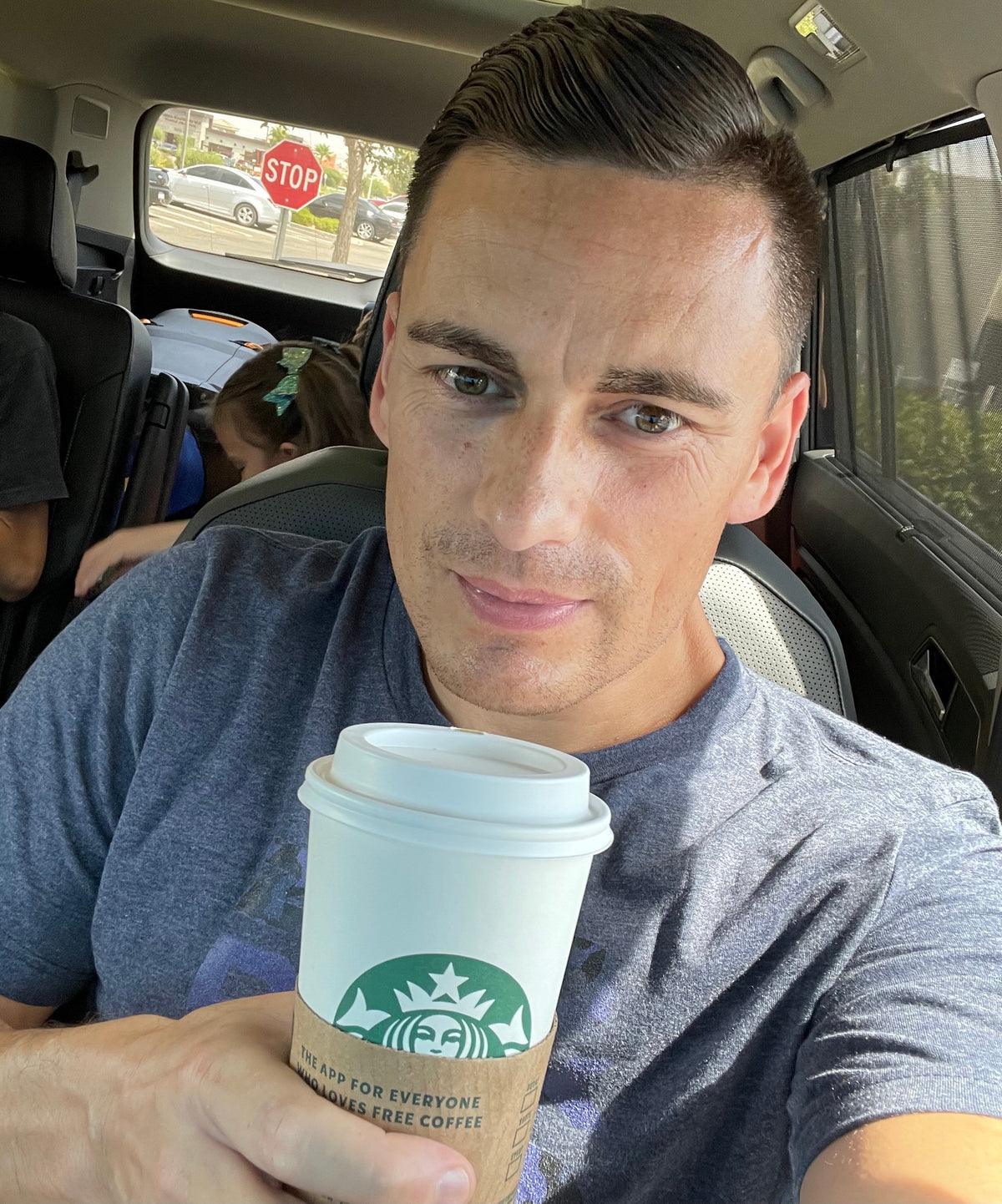A man in a car holds a Starbucks cup, wearing a blue t-shirt