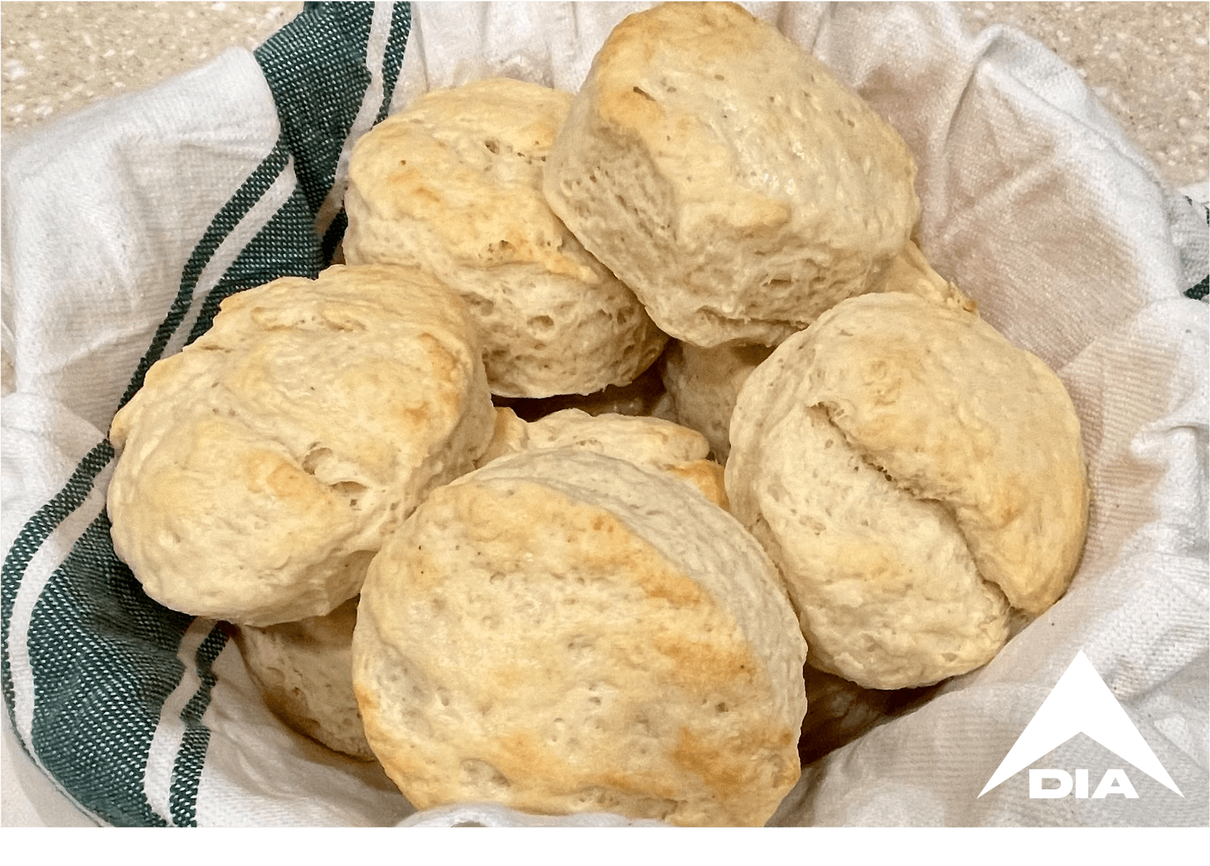 Basket of golden-brown biscuits on a green-striped towel in a kitchen, featuring a white DIA logo in the corner.
