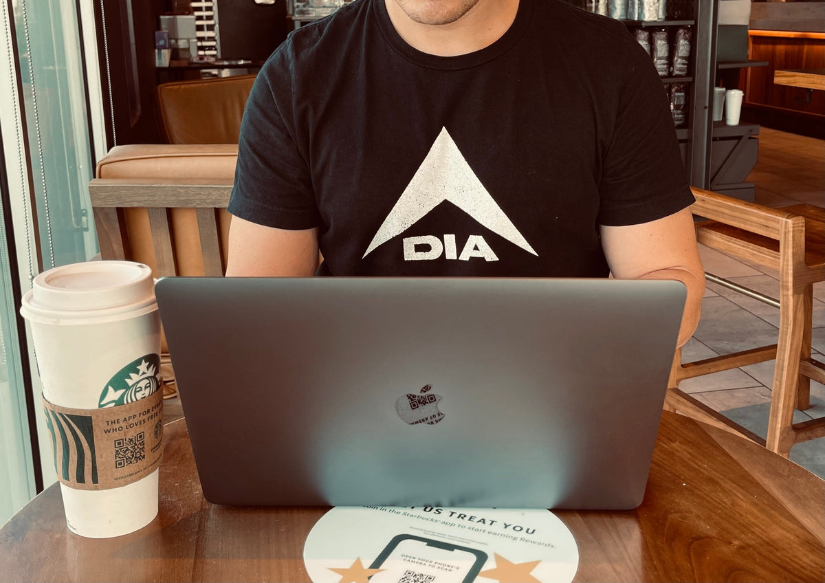 Man in a coffee shop working on a laptop with a Starbucks cup, wearing a black DIA logo t-shirt.