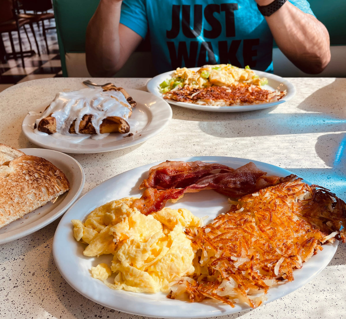 Man in a diner sits before plates of breakfast food, wearing a blue "JUST WAKE UP" DIA shirt.