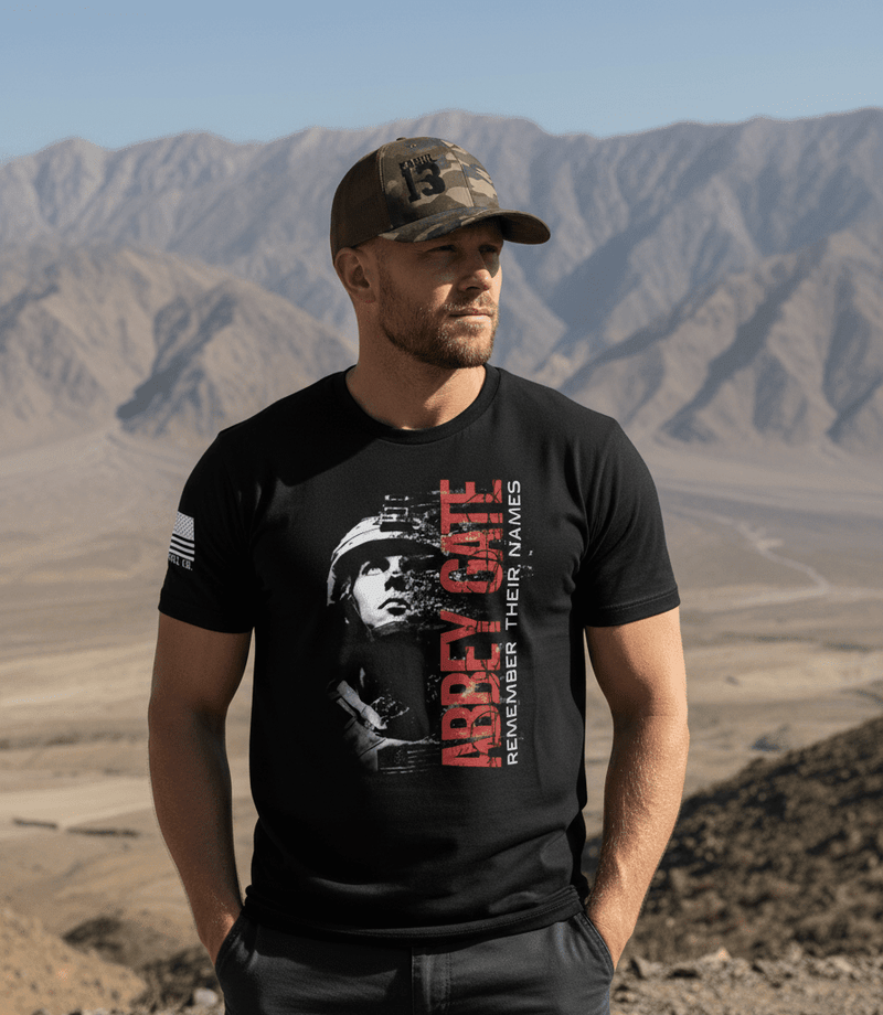 Man standing in a mountainous desert setting, wearing a camouflage "KABUL 13" trucker hat and an "ABBEY GATE" t-shirt.