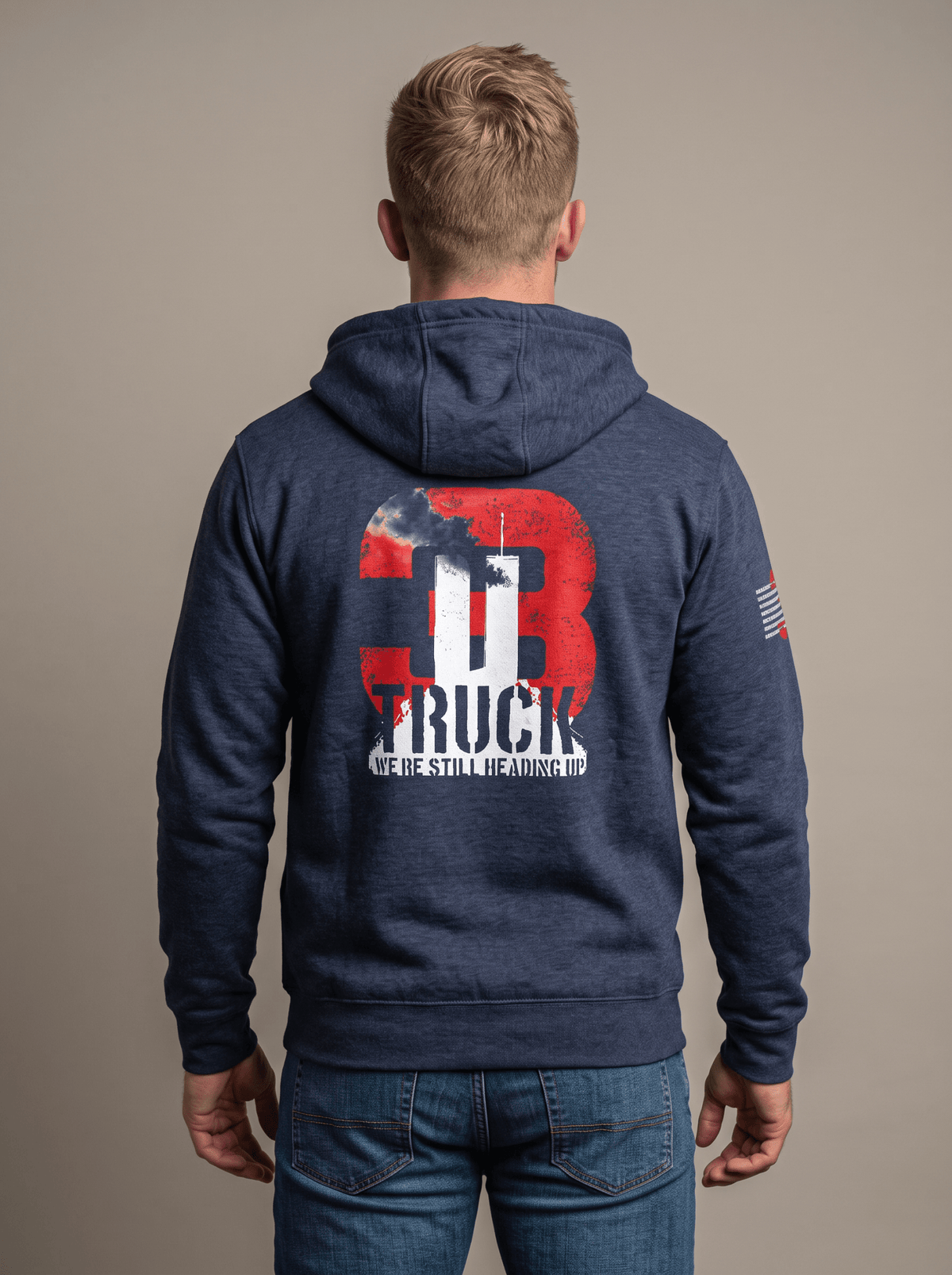 Man stands in studio facing away, wearing navy hoodie with "3 TRUCK WE'RE STILL HEADING UP" 9/11 graphic, American flag patch.   