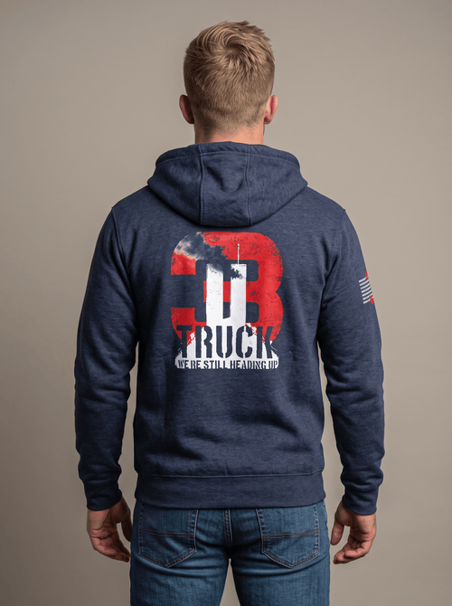 Man stands in studio facing away, wearing navy hoodie with "3 TRUCK WE'RE STILL HEADING UP" 9/11 graphic, American flag patch.   