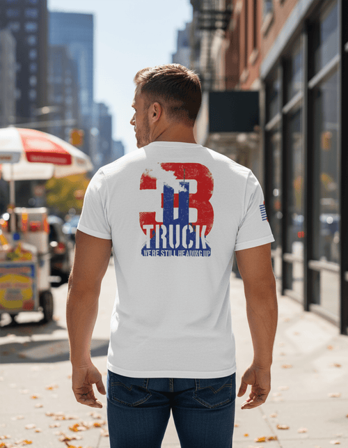 Man standing on a city street, back view of him wearing a white T-Shirt with red/blue "3 TRUCK" with twin towers graphic.