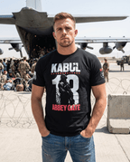 Man on an airfield, black Kabul 13 Abbey Gate shirt, standing close-up, front view with a military aircraft behind.