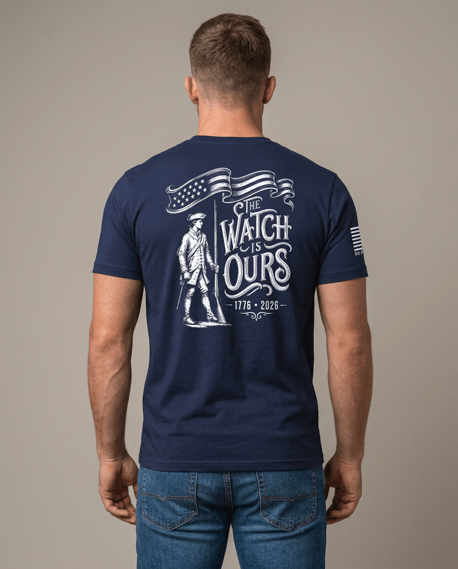 Man from behind in studio wearing navy t-shirt with "THE WATCH IS OURS 1776-2026" minuteman back graphic.