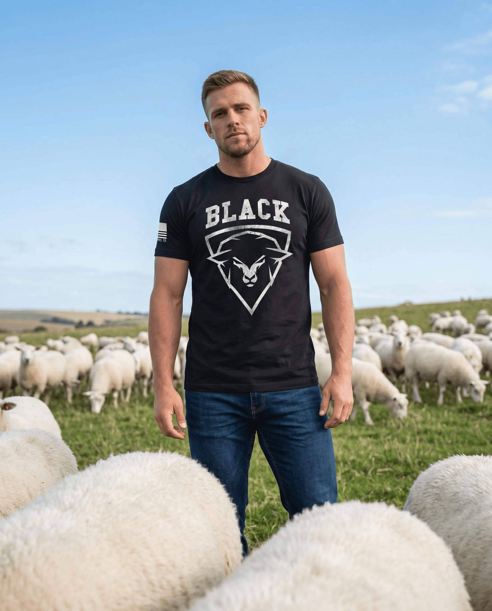 Man standing in a field of sheep, wearing a black t-shirt with a sheep graphic and "BLACK" text