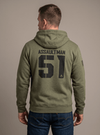 Man stands in a studio facing away, wearing a green hoodie with "ASSAULTMAN 51", DIA logo, and an American flag patch.
