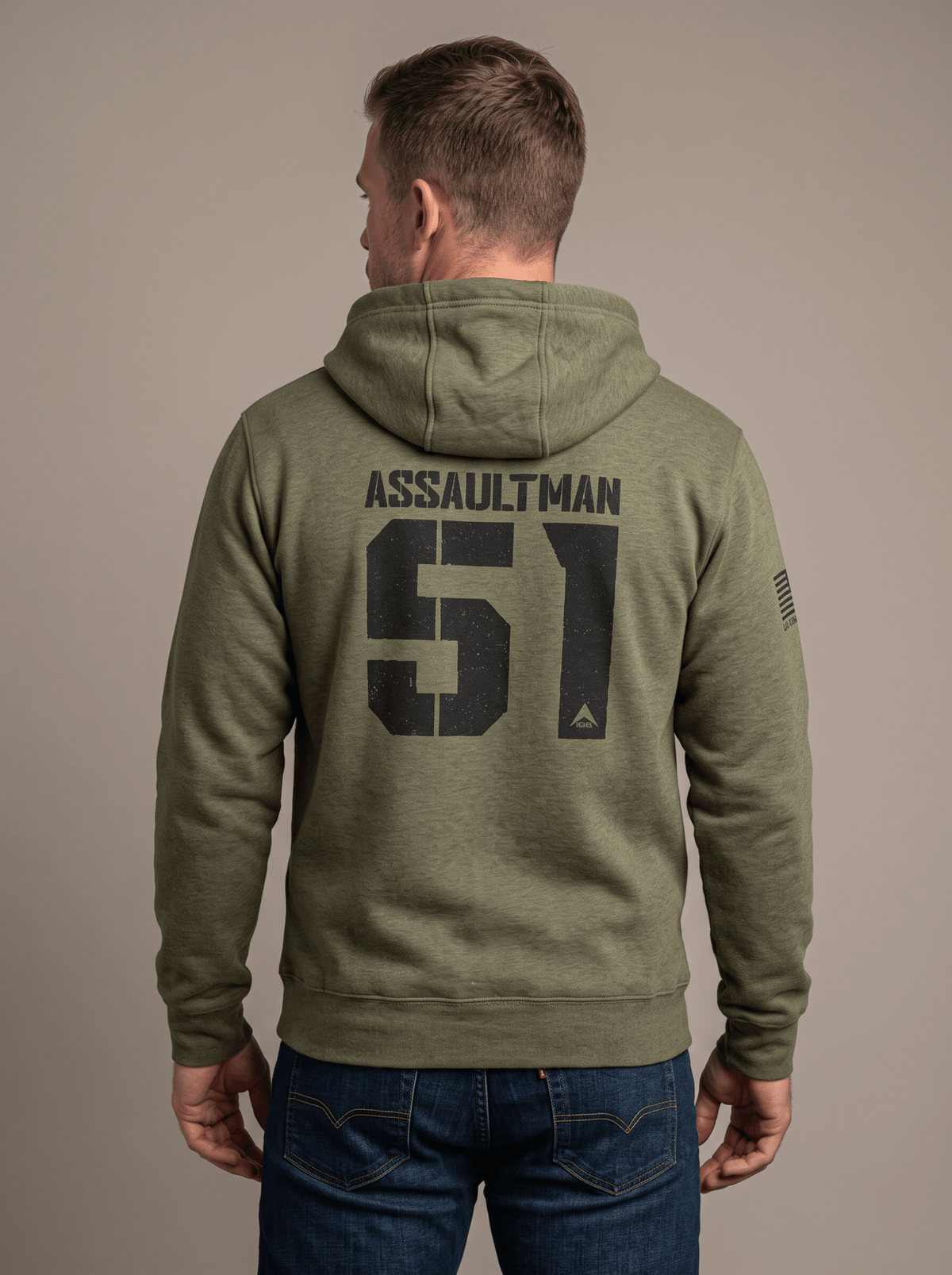 Man stands in a studio facing away, wearing a green hoodie with "ASSAULTMAN 51", DIA logo, and an American flag patch.