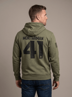 A man stands in a studio facing away, wearing a green hoodie with "MORTARMAN 41", a DIA logo, and an American flag patch.