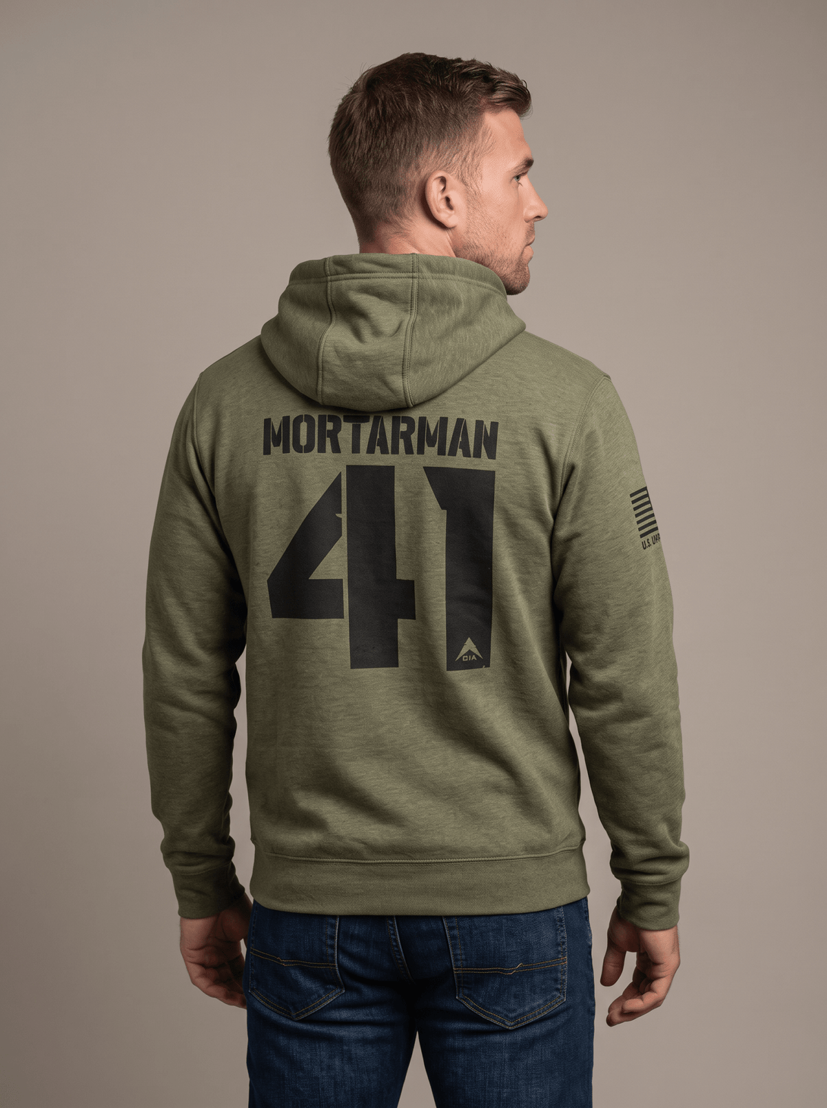 A man stands in a studio facing away, wearing a green hoodie with "MORTARMAN 41", a DIA logo, and an American flag patch.