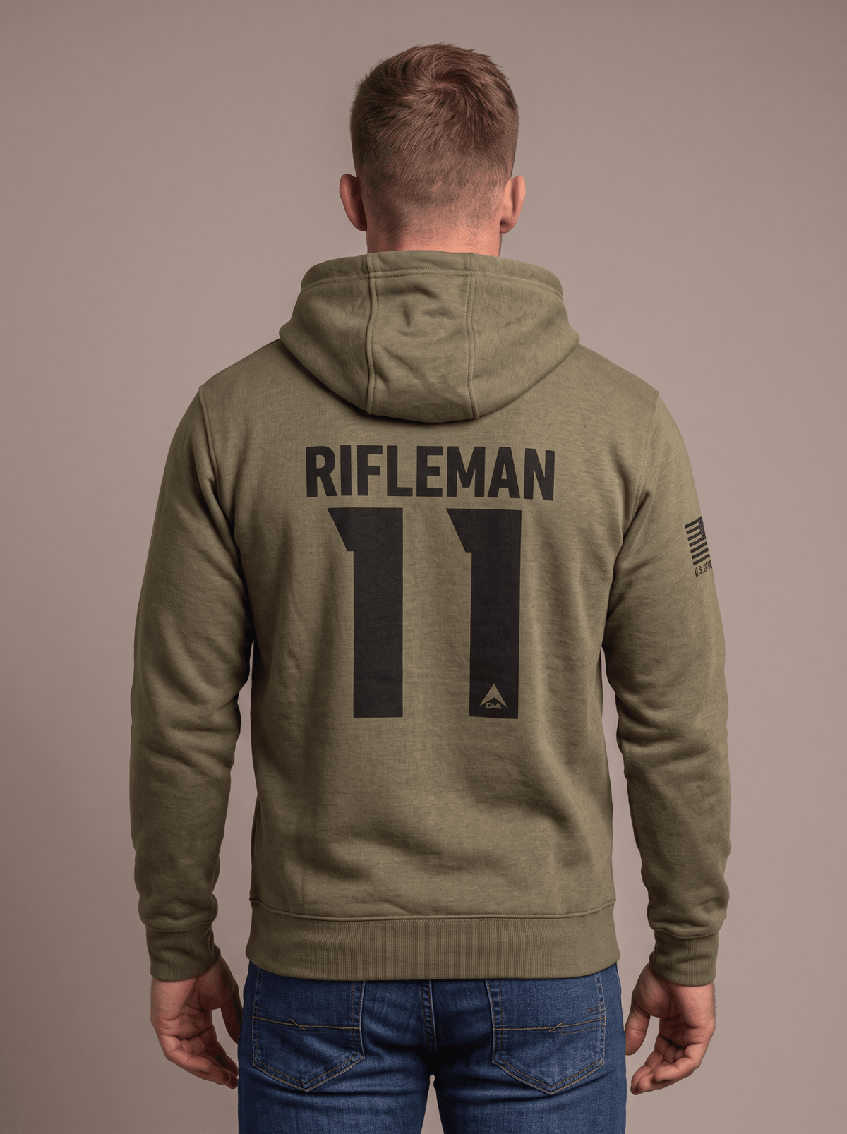 Man stands in studio facing away, wearing olive green hoodie with black "RIFLEMAN 11" back graphic and right sleeve American flag patch.