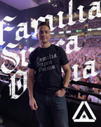A man stands in a crowded stadium wearing a black "Familia Supra Omnia" t-shirt and blue jeans.