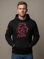 Man in studio wearing black hoodie featuring red "STAY FERAL" snarling raccoon graphic and American flag sleeve patch.