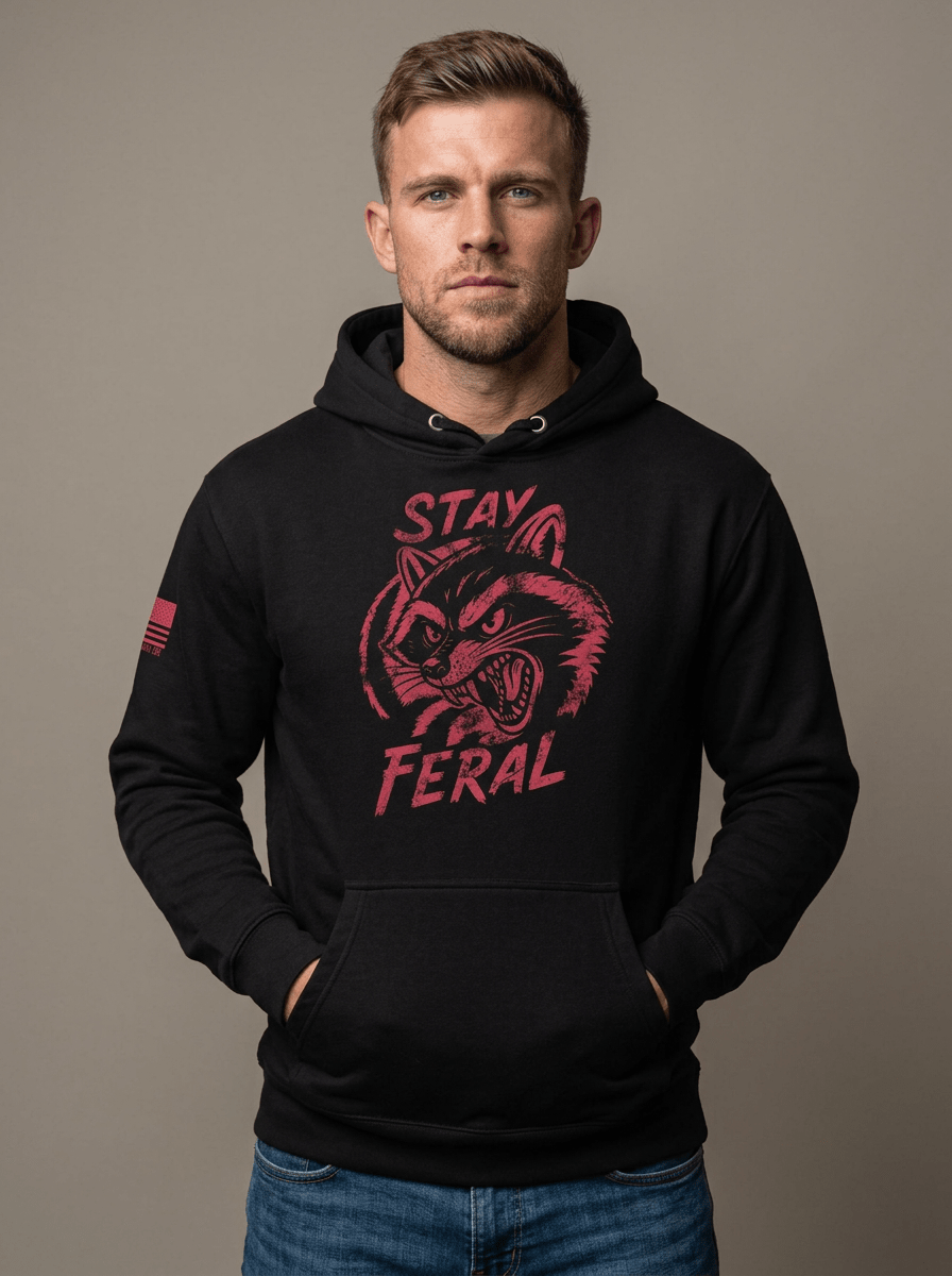 Man in studio wearing black hoodie featuring red "STAY FERAL" snarling raccoon graphic and American flag sleeve patch.