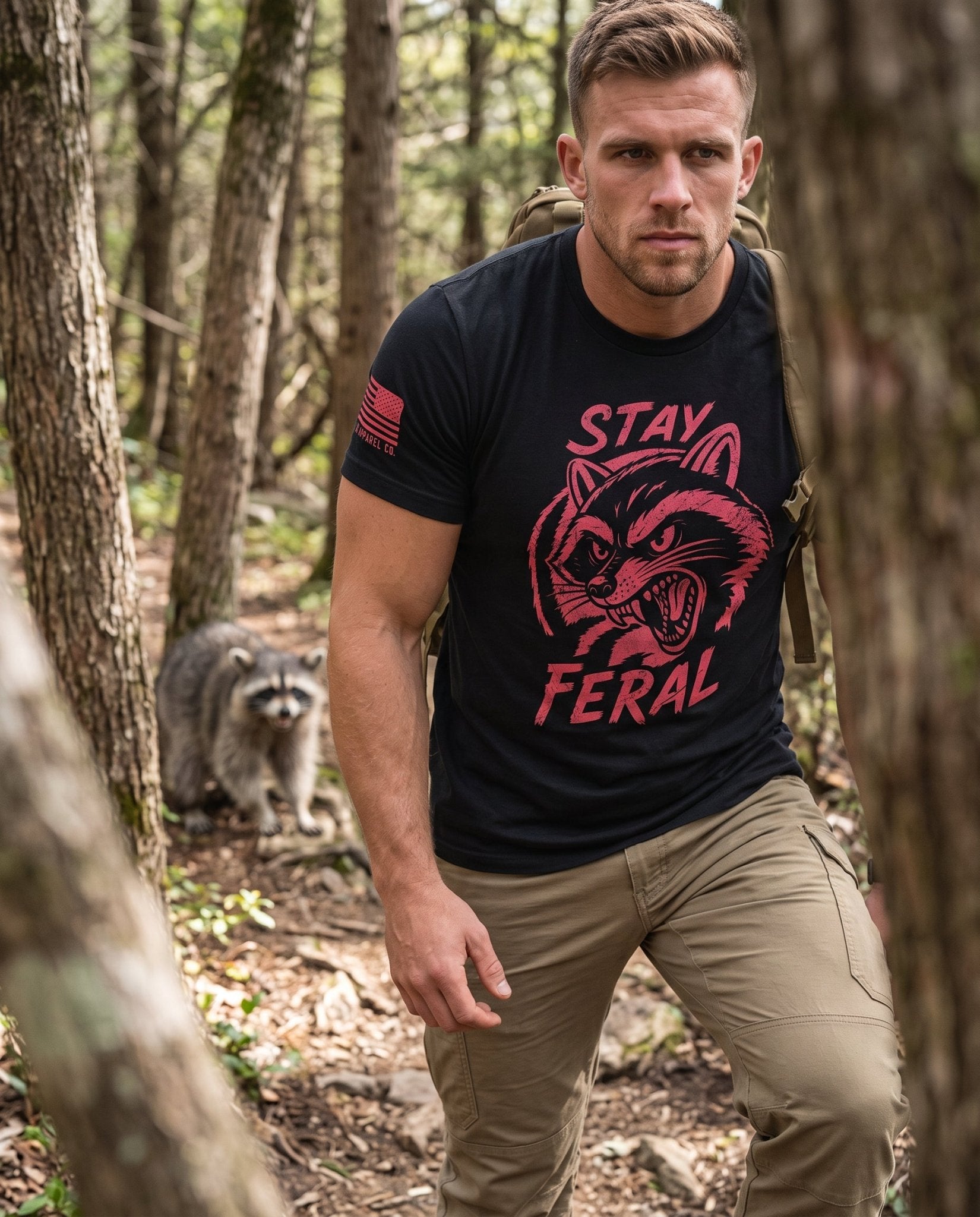 Man hiking in woods, wearing black "Stay Feral" raccoon t-shirt with American flag sleeve patch, raccoon behind.