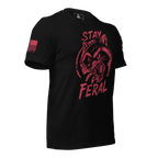 Black t-shirt shown angled right, with red "Stay Feral" snarling raccoon graphic and red American flag on right sleeve.