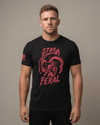 Man in studio wearing a black t-shirt with a red "STAY FERAL" snarling raccoon graphic and an American flag sleeve patch.