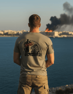 Man by a coastal city with smoke, wearing an olive green Benghazi shirt with an operator graphic and black American flag.