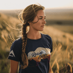 A woman stands in a grassy field at sunset wearing a navy blue t-shirt featuring a "Roam Free" buffalo graphic.