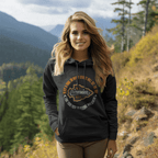 A woman stands in a sunny mountain setting, hands in her pockets, wearing a Dark Heather grey Pathfinder hoodie.