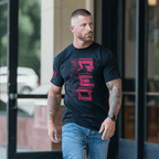 Man walking outside a glass door wearing a black USCG edition RED DIA shirt and blue jeans.