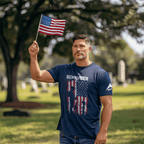Man, standing in a cemetery holding a flag, wearing a navy t-shirt with a soldier silhouette and a white DIA logo.
