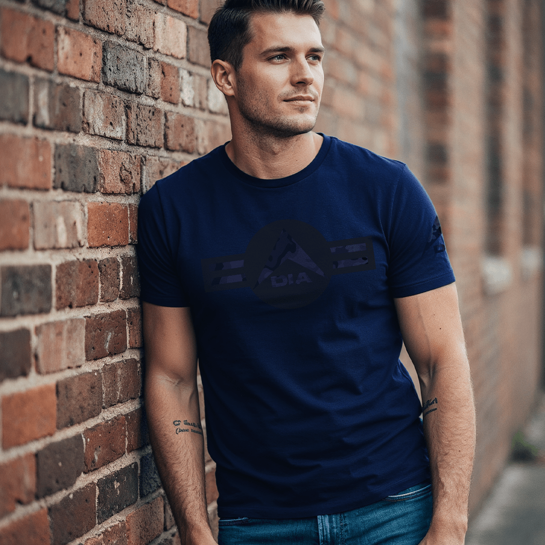 A man leans against a brick wall in an alley wearing a navy blue t-shirt with a tonal black DIA logo on the chest.