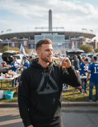 A man with a short, styled haircut and beard is smiling while drinking from a glass bottle at an outdoor tailgate party. He is standing in a crowded parking lot with a large stadium and a tall monument visible in the background. He is wearing a black hoodie featuring a large, tonal grey DIA logo on the chest and a subtle black American flag graphic on the right sleeve.