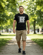 Man in black "THANK YOU FOR SERVING OVER THERE" t-shirt, khaki shorts, and sneakers walking on a park path.