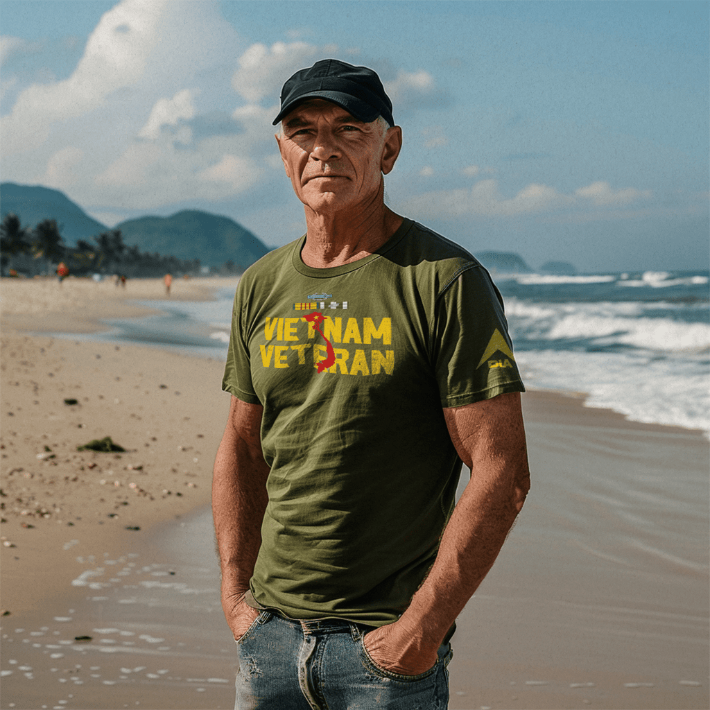 A man stands on a beach wearing an army green Vietnam Veteran t-shirt with a yellow DIA logo on the sleeve.