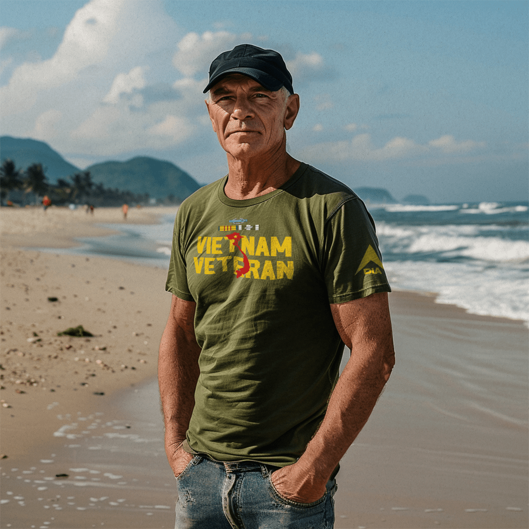 A man stands on a beach wearing an army green Vietnam Veteran t-shirt with a yellow DIA logo on the sleeve.