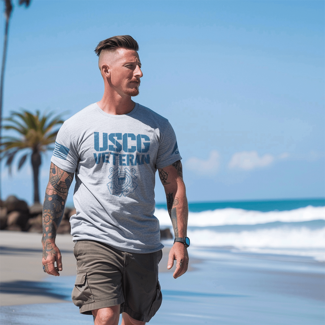 Man, walking on a sunny beach, wearing an athletic heather t-shirt with USCG Veteran text, Coast Guard emblem, and DIA logo.